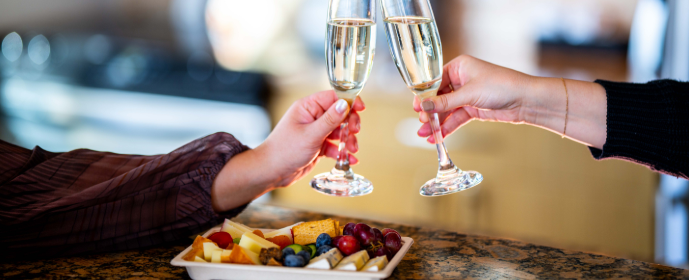 A Pair Of Hands Holding Wine Glasses Over A Plate Of Food